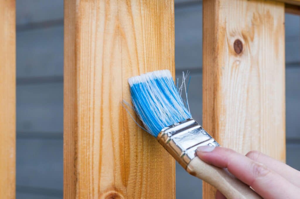 Application de lasure bois avec pinceau neuf sur balustrade A hand painting a wooden fence plank with a blue paintbrush outdoors. Perfect for DIY and renovation themes.