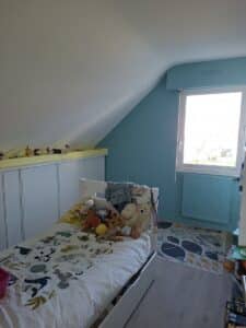 Chambre sous pente enfant. Mur turquoise, radiateur bleu. Lit blanc avec couette marine, peluches et étagère jaune.