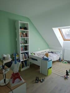 Chambre mansardée aux murs vert menthe, étagère blanche de livres et mangas, lit simple, et maquette Tour Eiffel sur le bureau.