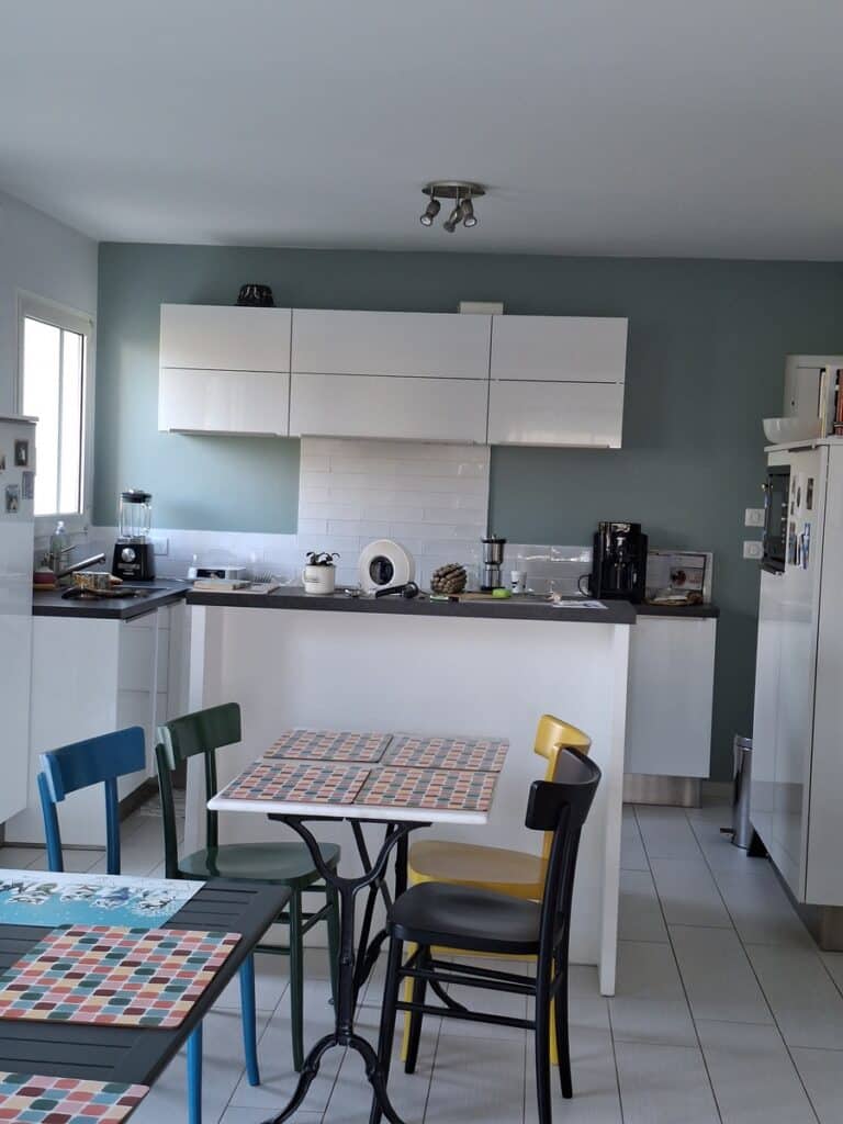Cuisine moderne blanche avec îlot et mur bleu canard. Chaises de salle à manger colorées (bleu, vert, jaune) sur carrelage gris clair.