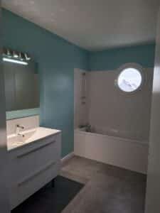 Salle de bain moderne et lumineuse. Mur bleu sarcelle, meuble vasque blanc, baignoire carrelée et fenêtre ronde style hublot.
