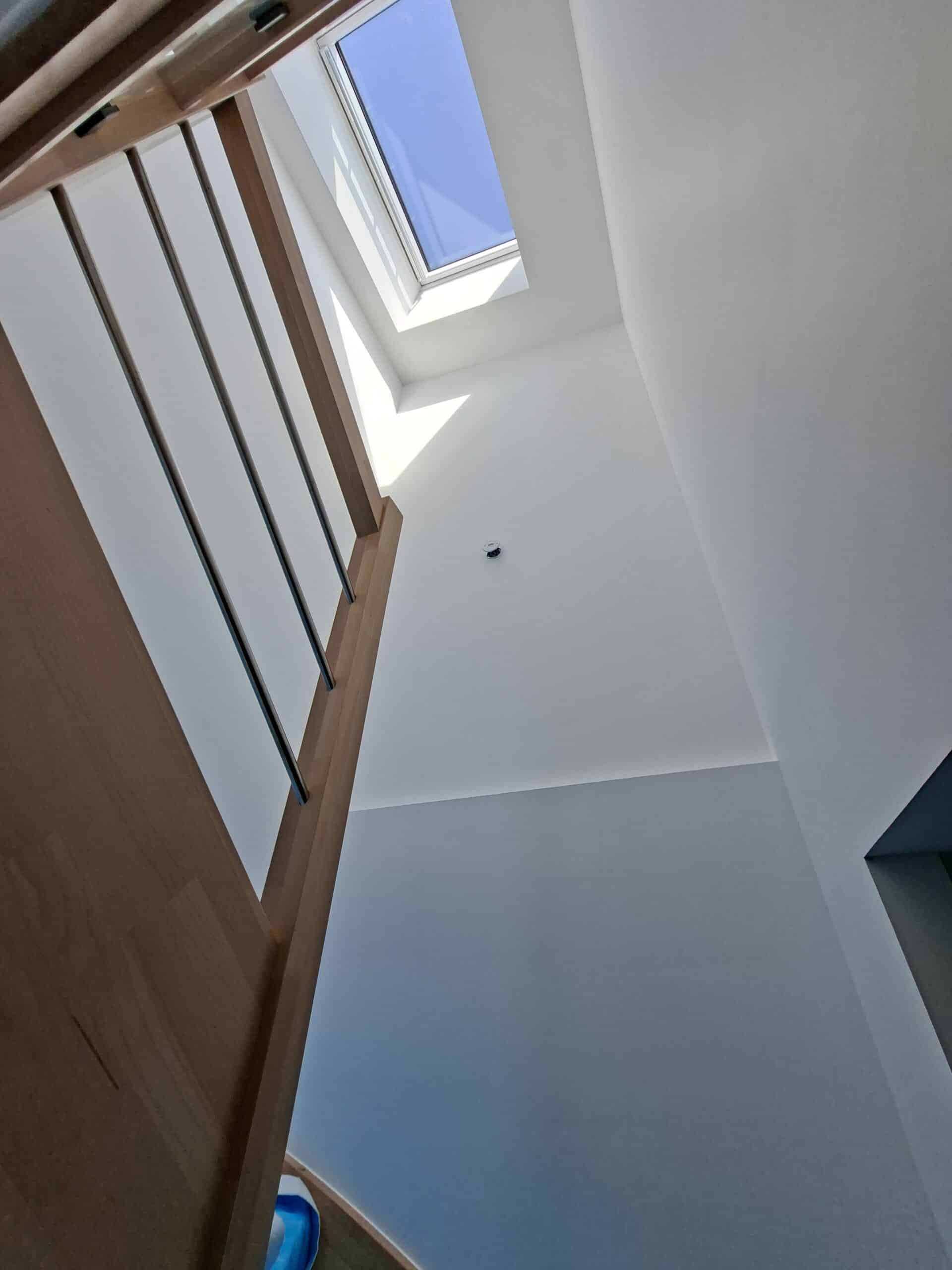 Escalier en bois et garde-corps métal sous un puits de lumière offrant une clarté naturelle et vue sur le ciel bleu.