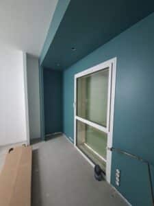 Intérieur en rénovation avec un mur bleu canard, une fenêtre blanche et un sol en béton brut.