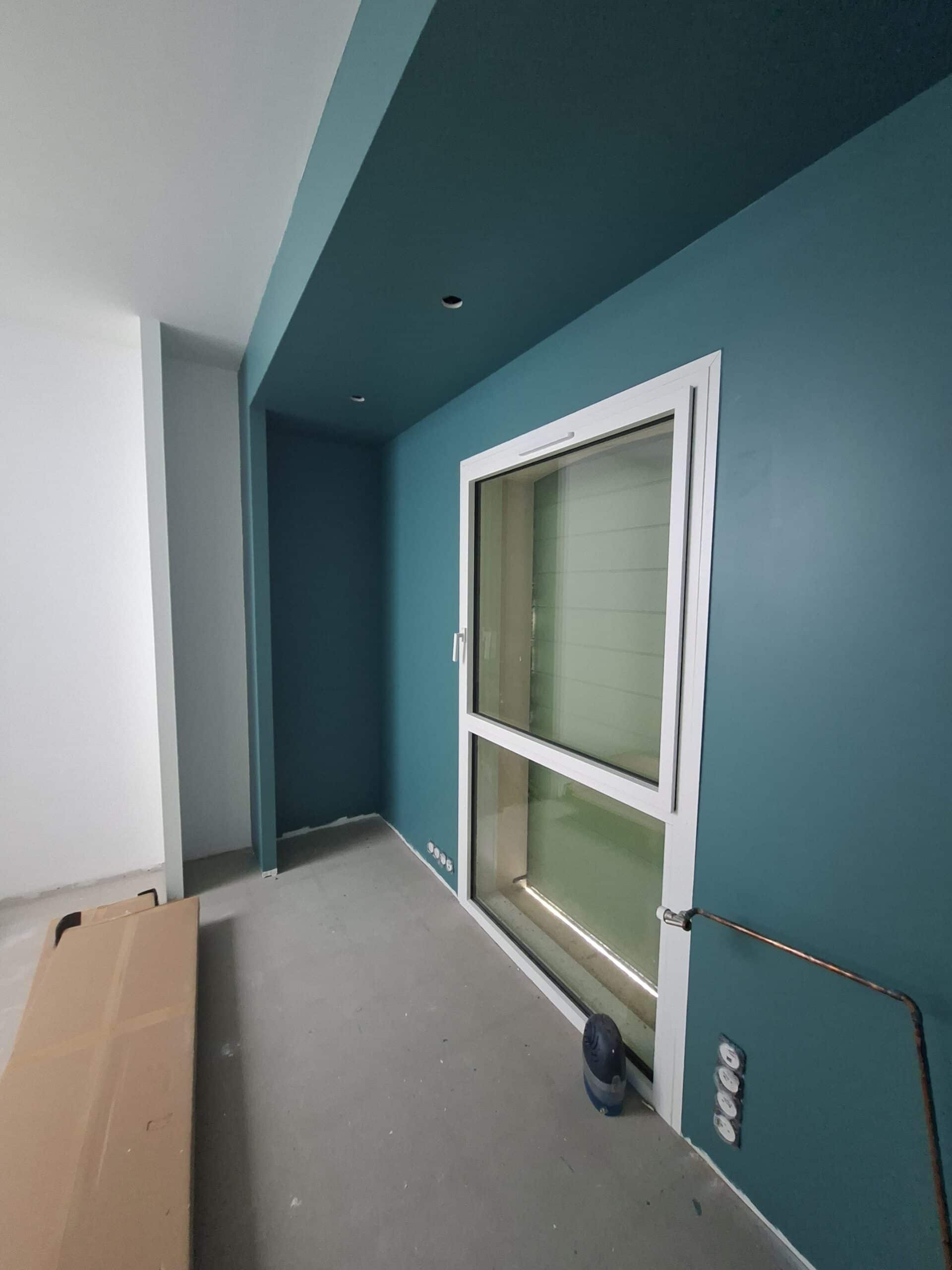 Intérieur en rénovation avec un mur bleu canard, une fenêtre blanche et un sol en béton brut.