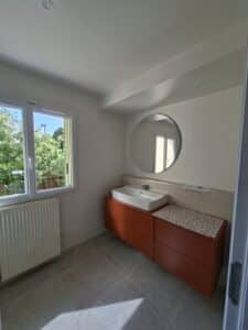 Salle de bain moderne avec meuble vasque terracotta, évier blanc rectangulaire, miroir rond et grande fenêtre lumineuse.