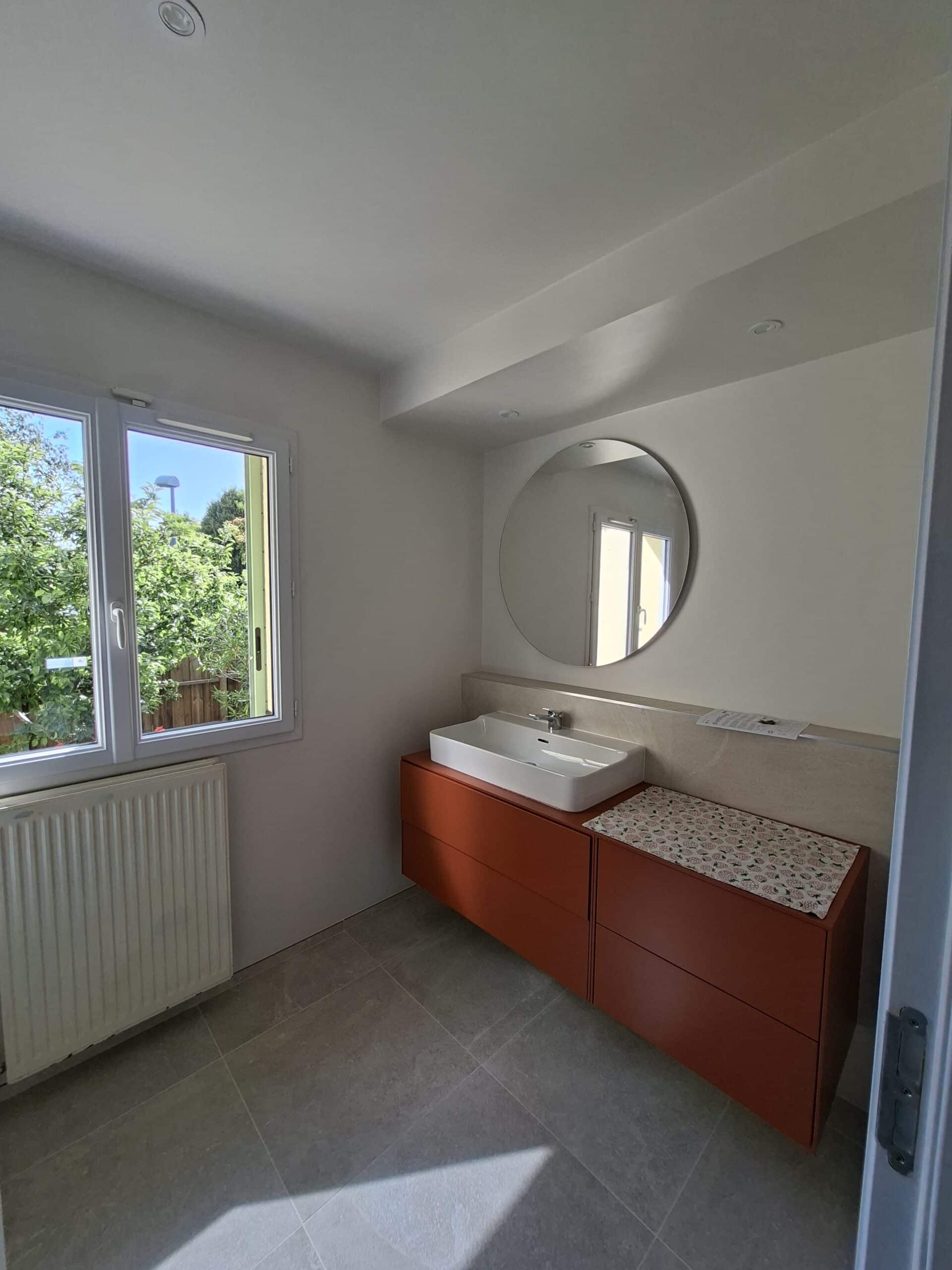 Salle de bain moderne avec meuble vasque terracotta, évier blanc rectangulaire, miroir rond et grande fenêtre lumineuse.
